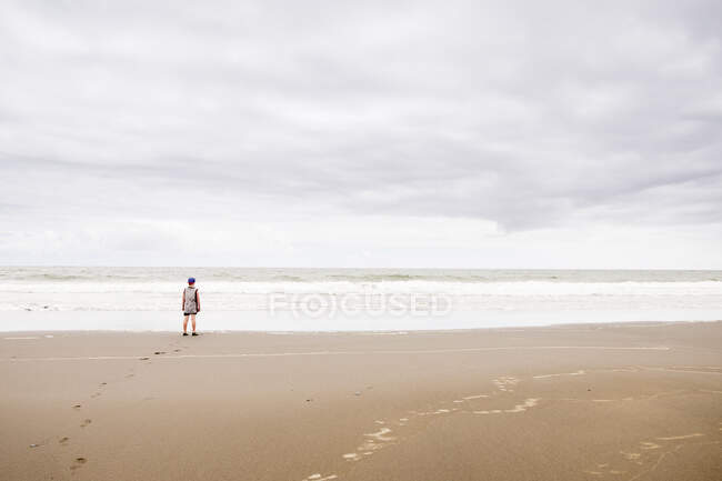 Jeune garçon debout sur la plage regardant l'eau — Photo de stock