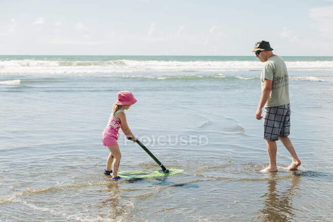 Chica joven y su padre jugando en la playa con un tablero de skim - foto de stock