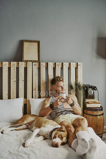 Un ragazzo biondo con una tazza di caffè e il suo cane a letto. Stile di vita concetto — Foto stock