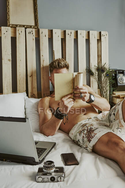 Ein Mann liest ein Buch, während er in einem Hotelbett mit Handy und Laptop arbeitet. Entspannungskonzept — Stockfoto