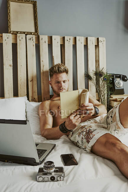 Ein Mann liest ein Buch, während er in einem Hotelbett mit Handy und Laptop arbeitet. Entspannungskonzept — Stockfoto