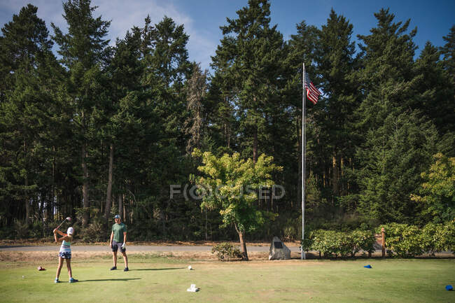 Chica joven en el golf tee balanceo club de golf, mientras que el hombre mira en día soleado - foto de stock