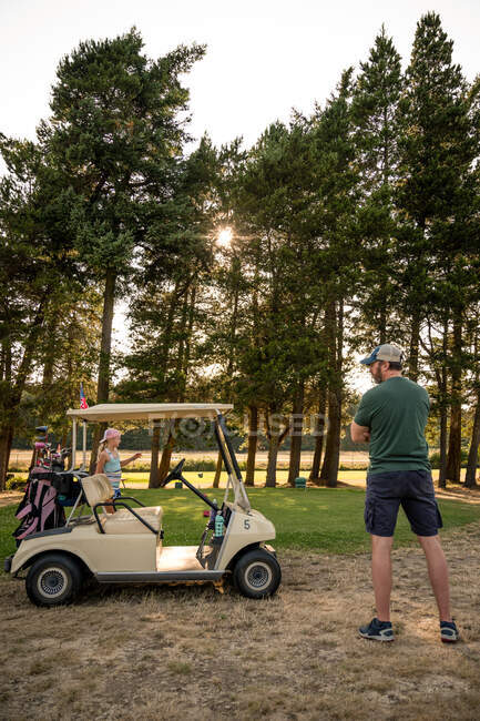 Chica joven con cola de caballo rubia y hombre de mediana edad de pie en el campo de golf con carrito de golf en el día soleado. - foto de stock