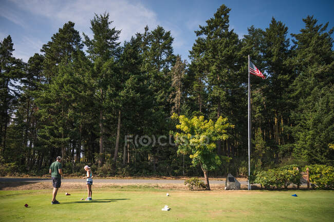 Man and young girl at golf tee with american flag and trees in background — Stock Photo
