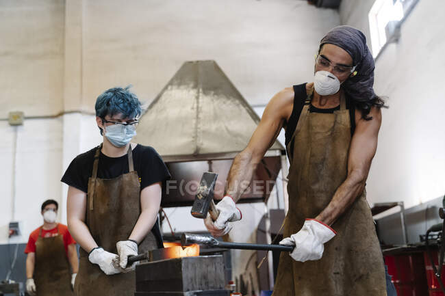 Blacksmiths in masks hitting hot metal with hammer while working — Stock Photo
