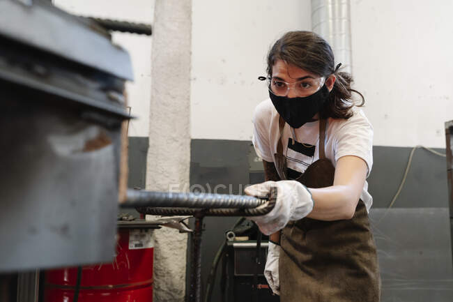 Жінка-коваль в захисній масці в майстерні виконує металеві роботи — стокове фото