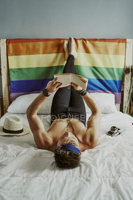 Молодий чоловік у ліжку, який читає книгу з гей-прапором за спиною.. — стокове фото