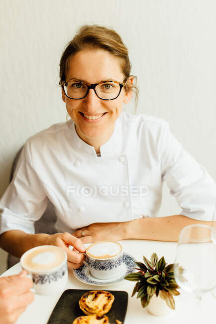 Donna in uniforme bianca sorridente seduta a tavola con cappuccino — Foto stock