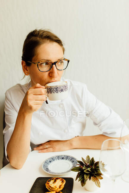 Жінка в білій уніформі п'є капучино за столом в кафе — стокове фото