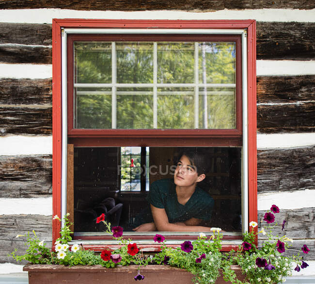 Adolescente chico mirando por la ventana de rústico cabaña de madera en el día de verano. - foto de stock