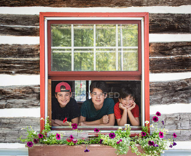 Tres chicos mirando por la ventana de la cabaña de madera rústica en el día de verano. - foto de stock