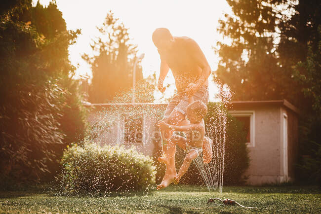 Отец и ребенок прыгают через воду из разбрызгивателя в саду — стоковое фото
