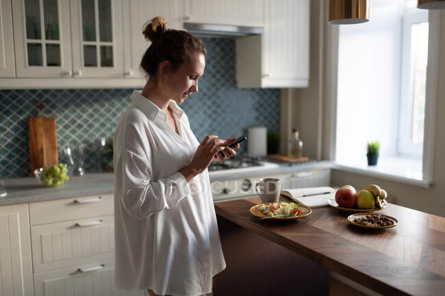 Side view female using smartphone while having breakfast before work at home — Stock Photo
