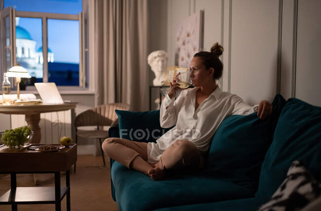 Доросла жінка сидить на дивані і п'є вино під час відпочинку після роботи вдома — стокове фото