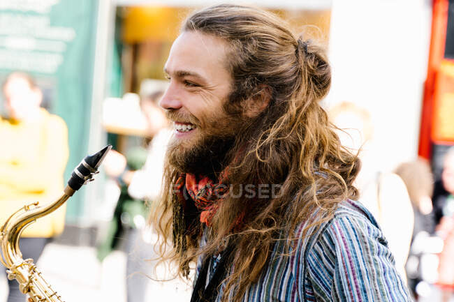 Retrato del perfil de un joven con el pelo largo sonriendo mientras toca el saxofón en la calle - foto de stock