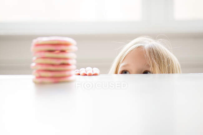 Ragazza bionda sbirciando pila di biscotti di zucchero glassato rosa sul tavolo — Foto stock