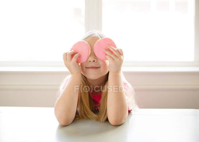 Cute blond girl holding pink frosted sugar cookies over eyes — Stock Photo