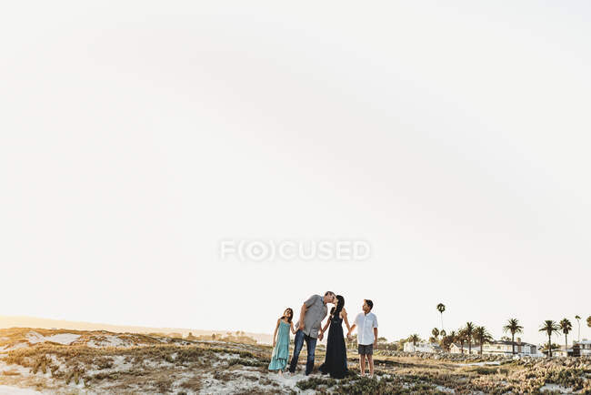 Familia cogida de la mano en la playa como papá besa a mamá - foto de stock
