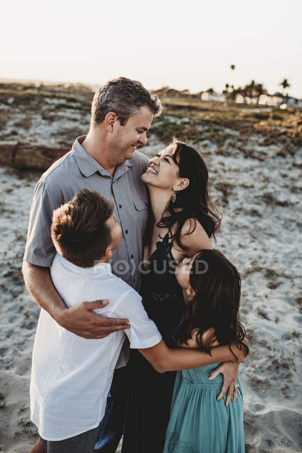 Семья обнимается на песчаной дюне с улыбающимися родителями и двумя детьми — стоковое фото