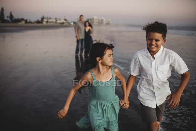 Riéndose niño preadolescente y hermana corriendo por delante de los padres en la playa - foto de stock