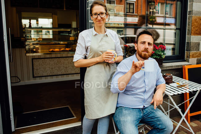 Man and woman smiling at camera while drinking tea at entrance of cafe — Stock Photo