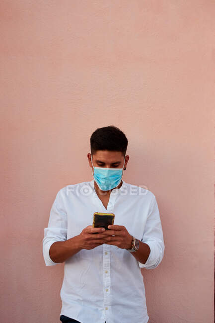 Молодий латинський чоловік з маскою дивиться свій телефон на рожевому фоні — стокове фото