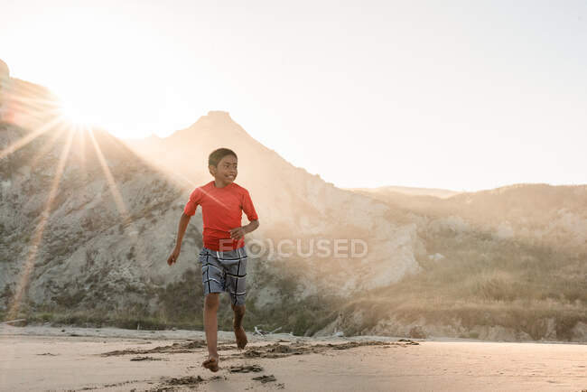 Latino tween menino correndo na praia com montanhas no fundo — Fotografia de Stock