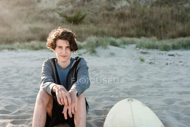 Adolescente de pelo largo sentado junto a la tabla de surf en una playa en Nueva Zelanda - foto de stock