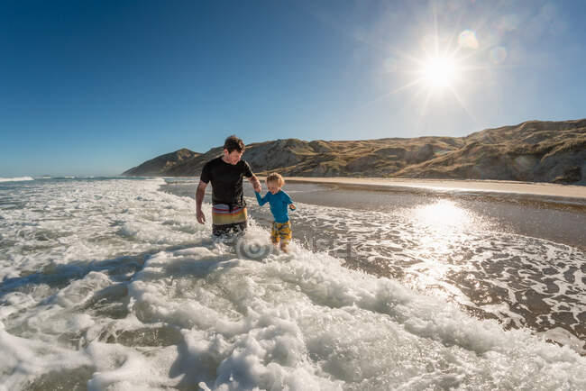 Père et enfant souriants jouant en vagues par une journée ensoleillée sur une plage en Nouvelle-Zélande — Photo de stock
