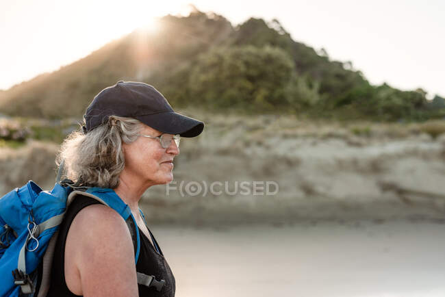 Femme âgée active portant un sac à dos et un chapeau marchant sur une plage en Nouvelle-Zélande — Photo de stock