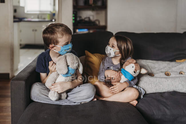 Niña en edad preescolar y niño en edad escolar con máscaras sentadas en el sofá - foto de stock