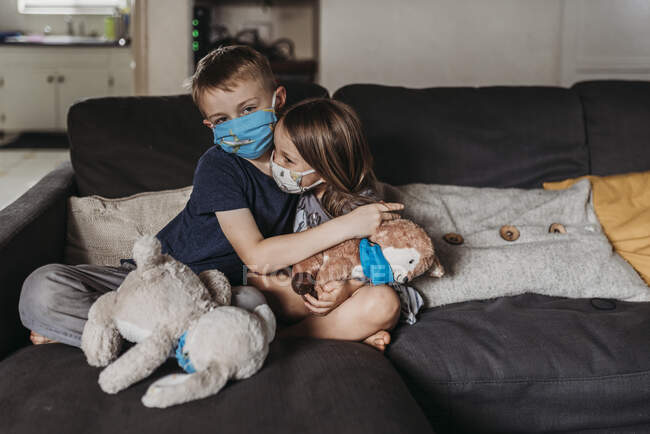 Chica joven y niño en edad escolar con máscaras abrazando y sonriendo en el sofá - foto de stock