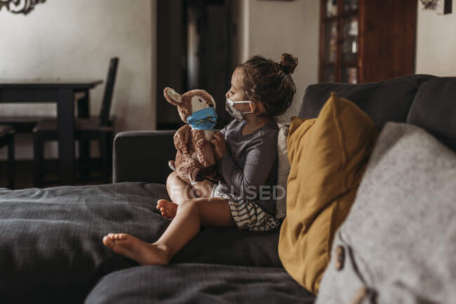 Side view of preschool age girl with mask on cuddling masked toy — Stock Photo