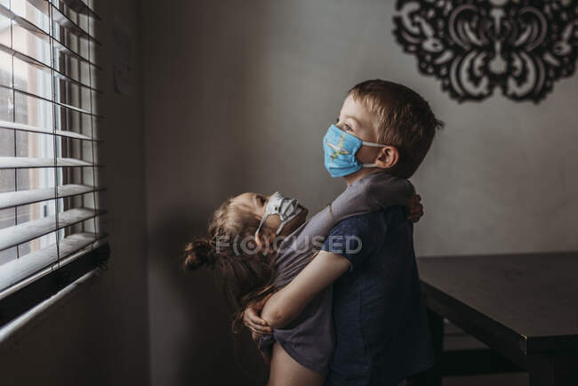 Retrato de estilo de vida de hermanos jóvenes con máscaras en abrazos en casa - foto de stock