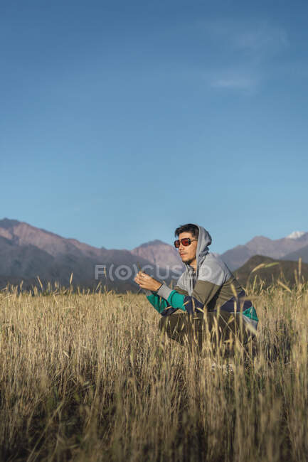 Young man with sunglasses sitting on in field looking at mountains wit — Stock Photo