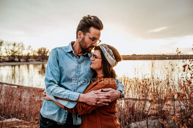 Hüftiges junges Paar, das sich an einem warmen Herbstabend an einem See umarmt — Stockfoto