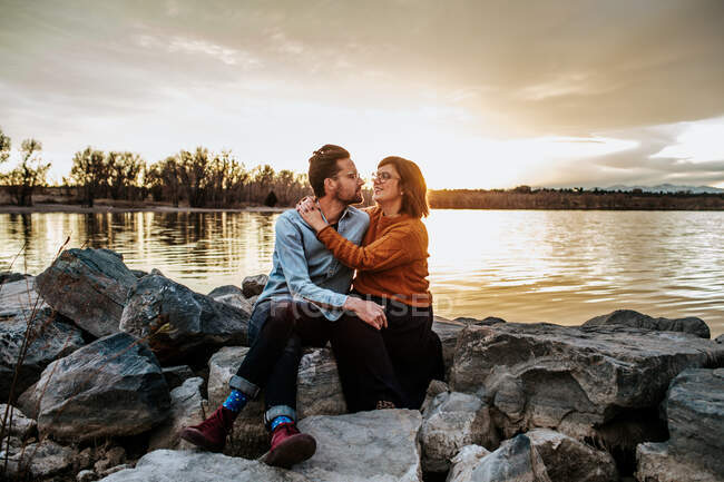 Esposo y esposa sentados en las rocas cerca de un lago en una noche de otoño - foto de stock