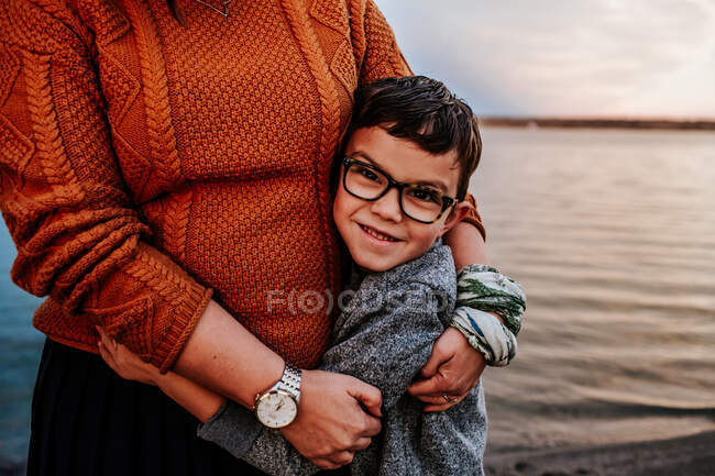 Щасливий молодий хлопчик обіймає маму біля озера в осінній вечір — стокове фото