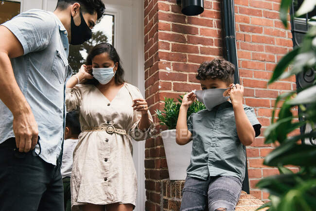 Familia e hijo pequeño que se pone máscaras para salir de casa de piedra rojiza - foto de stock