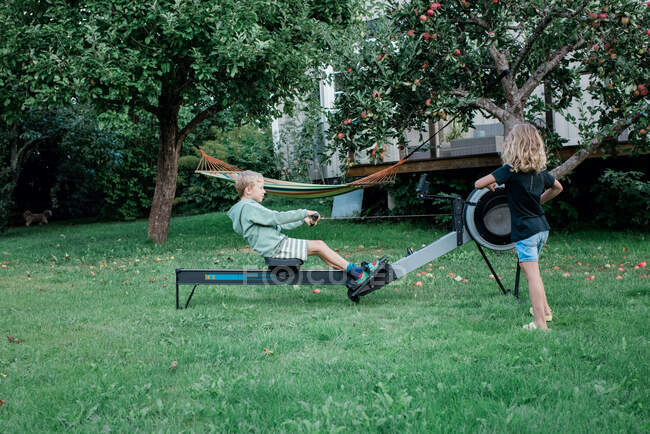 Брат и сестра с помощью домашнего тренажерного зала гребная машина в своем саду — стоковое фото
