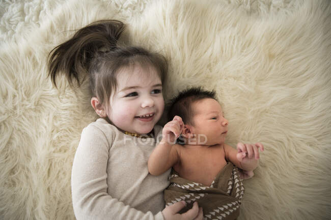 Старшая сестра обнимает новорожденного брата, пока он лежит на пушистом ковре — стоковое фото