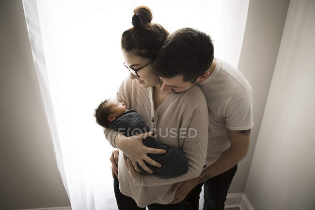 Los padres de la generación Z se acurrucan por la ventana y admiran a su bebé recién nacido - foto de stock