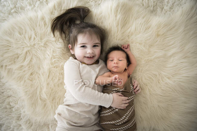 Happy Toddler Girl acurruca al hermano recién nacido, acostándose sobre una alfombra blanca difusa - foto de stock