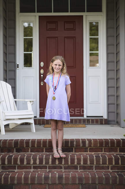 Fille blonde souriante portant une médaille debout sur le porche avant en brique — Photo de stock