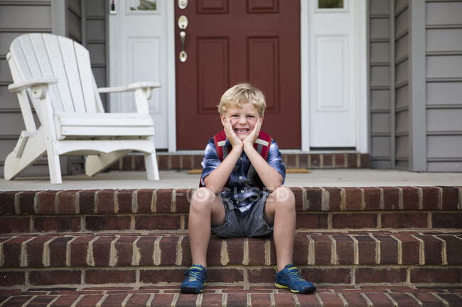 Sourire blonde garçon assis sur brique devant marches avec tête dans les mains — Photo de stock
