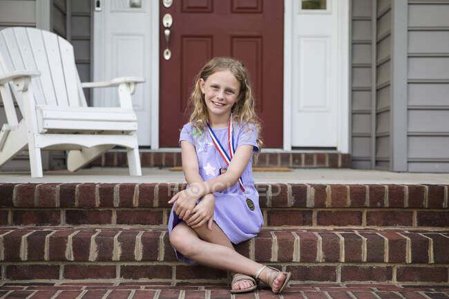Sonriente chica rubia con una medalla se sienta en frente de ladrillo pasos delanteros - foto de stock
