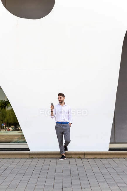 Молодий бородатий чоловік спирається на білу стіну за допомогою мобільного телефону — стокове фото