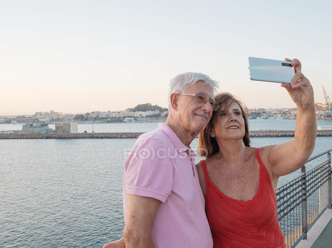 Happy elderly couple taking selfie photo on smart phone while sitting on the bridge in the city. — Stock Photo