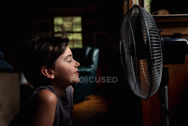 Niño sonriendo y refrescándose delante de un ventilador en habitación oscura - foto de stock