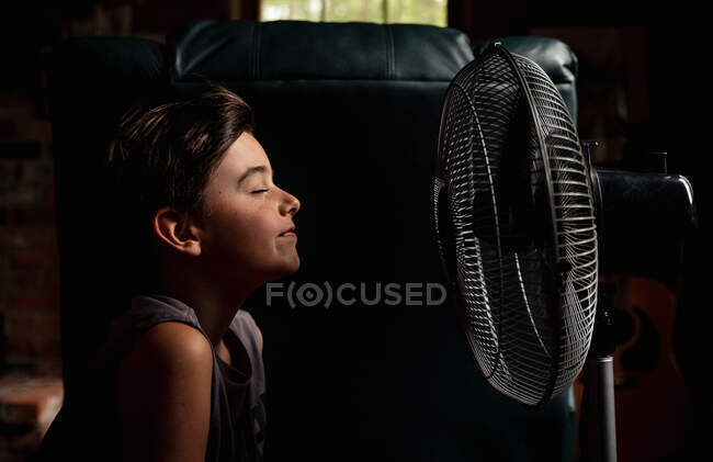 Kleiner Junge mit geschlossenen Augen kühlt sich vor einem Ventilator im dunklen Raum ab — Stockfoto